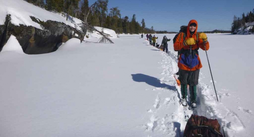 a line of cross country skiers, each pulling small sleds, make their way across a frozen, snowy lake